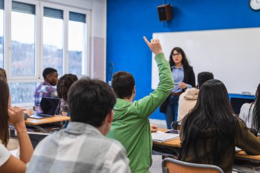 Lise öğrencisi ders sırasında soru sormak için elini kaldırırken, öğretmen ve sınıf arkadaşları arka planda