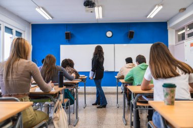 Lise öğrencileri sınıfta notlar alırken öğretmenleri beyaz tahtanın yanında duran dersi anlatıyor.