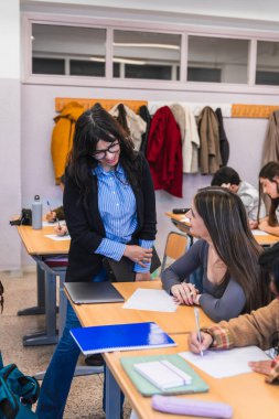 Gülümseyen bayan öğretmen bir sınıf sınavında lise öğrencisine rehberlik ediyor, destekleyici ve odaklanmış bir öğrenme ortamı sağlıyor.