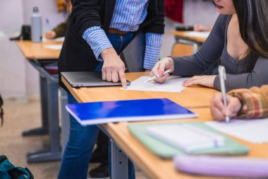 Sınav kağıdına işaret eden öğretmen, bir lise öğrencisine odaklanmış bir sınıf ortamında sınav sırasında rehberlik ediyor.