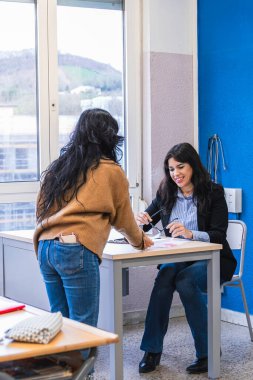 Lise öğrencisi, öğretmenine ödev teslim ediyor. Sınıfta, masasında oturuyor.