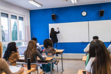 Lise öğretmeni beyaz tahtaya yazı yazarken sınıftaki çeşitli öğrenci gruplarına ders veriyor.