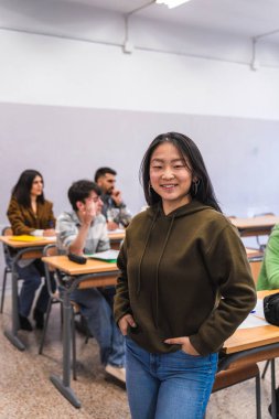 Sınıf arkadaşları ders çalışırken elleri ceplerinde duran gülümseyen Asyalı bir lise öğrencisinin portresi.