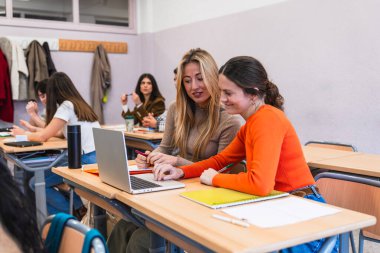 İki lise öğrencisi gülümsüyor ve sınıfta bir dizüstü bilgisayarda birlikte çalışırken, diğer öğrenciler arka planda öğreniyor.