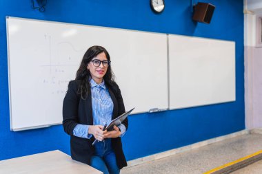 Lise öğretmeni elinde bir not defteri tutuyor ve sınıfta beyaz tahta ve matematik denklemleriyle gülümsüyor.