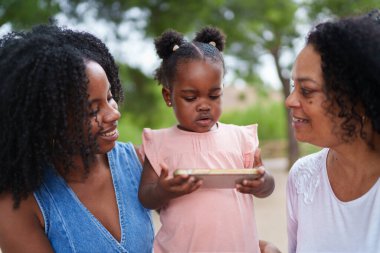 Küçük çocuk, annesi ve büyükannesiyle akıllı telefon kullanıyor. Parkta ailecek vakit geçiriyorlar.