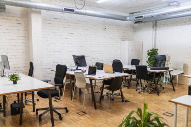 Masaların üzerinde beyaz tuğlalı duvar, ahşap zemin, dizüstü bilgisayarlar ve bilgisayarlar olan modern bir çalışma alanı.