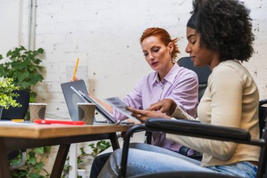 İki genç iş kadını, biri tekerlekli sandalye kullanıyor, modern ortak çalışma alanında bir proje üzerinde çalışıyorlar, belgeleri gözden geçiriyorlar ve bir dizüstü bilgisayar kullanıyorlar.