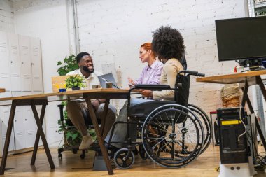 İki iş arkadaşı, tekerlekli sandalye kullanıyor, modern ofiste proje üzerinde işbirliği yapıyor, işyeri kapsayıcılığını ve takım çalışmasını gösteriyor