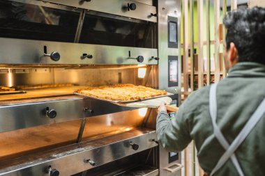 Baker profesyonel bir fırından bir tepsi altın esmer ekmek alıyor, ticari bir fırında pişirme sürecini gösteriyor.