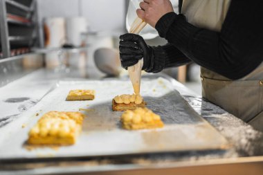 Profesyonel bir fırıncı boru torbasıyla hamur işlerine krema ekliyor, fırın mutfağında metal bir masa üzerinde çalışıyor.
