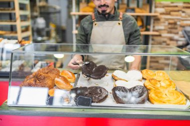 Baker çikolata kaplı palmieri cam bir vitrine koymak için maşa kullanıyor, arkasında çeşitli taze hamur işleri var.