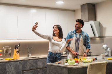 Genç çift, modern mutfaklarında sağlıklı bir salata hazırlarken akıllı telefonlarıyla selfie çekerek eğleniyor.