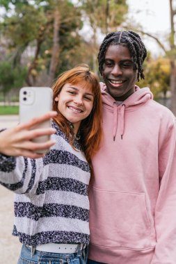 Genç, çok ırklı bir çift, ağaçlar ve doğanın güzelliği ile çevrili canlı bir parkta gülümseyip selfie çekiyorlar.