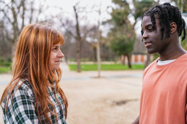 Genç, çok ırklı bir çift, neşe dolu bir sohbetin tadını çıkarıyor ve birlikte yeşil bir parkta rahatlarken gülücüklerini paylaşıyor.