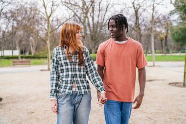 Gündelik giysiler içinde genç bir çift el ele tutuşup parkta yürüyor ve romantik bir gezinti yapıyorlar.