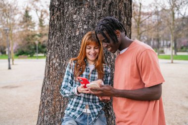 Parkta ağaç gövdesine yaslanırken akıllı telefonlar kullanan ve gülümseyen çok ırklı mutlu çift.