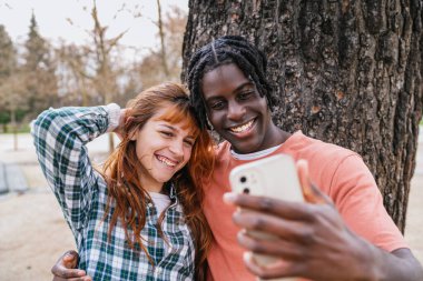Parkta akıllı bir telefonla selfie çeken, mutlu bir anın tadını çıkaran, çok kültürlü bir çift.