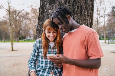 Parkta ağaca yaslanan mutlu çoklu etnik çift, birlikte akıllı telefon kullanırken gülümsüyor ve birlikte vakit geçiriyorlar.