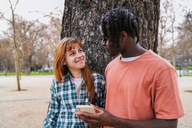 Genç, çok ırklı bir çift gülümsüyor ve akıllı bir telefon kullanıyor. Bu arada yeşil alanlarla dolu canlı bir parkta birlikteliklerinin tadını çıkarıyorlar.