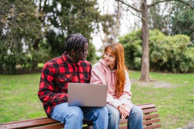 Parktaki bankta dizüstü bilgisayar kullanan ve birbirlerine bakıp gülümseyen çok kültürlü genç çift.