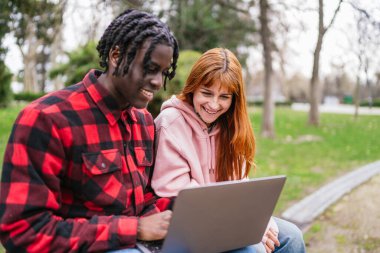 Dizüstü bilgisayar kullanan, parkta bankta oturan, ekrana bakan ve gülümseyen mutlu ve çok ırklı genç çift.