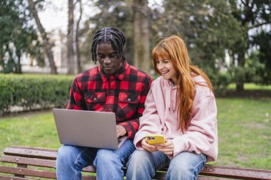 Genç, çok ırklı bir çift, parktaki bankta oturuyor. Çevrelerindeki doğanın güzelliğinin tadını çıkarırken dizüstü bilgisayar ve akıllı telefon kullanıyorlar.