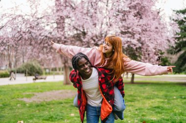 Mutlu çoklu etnik çiftler, çiçek açan parkta bahar gününün tadını çıkarıyorlar, erkek arkadaşı sırtında kız arkadaşını taşıyor, uçak taklidi yapıyor.