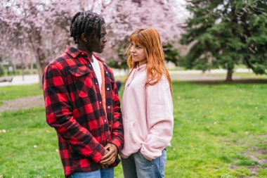 Genç, çok ırklı bir çift arka planda çiçek açan ağaçlarla parkta eğleniyor.