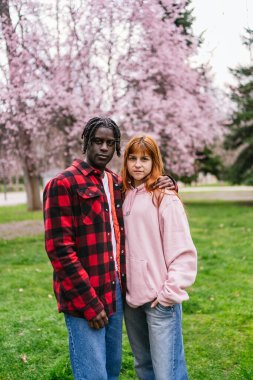 Arka planda kiraz çiçekleri olan parkta kucaklaşan çok ırklı genç çift, aşkı ve çeşitliliği kutluyorlar.