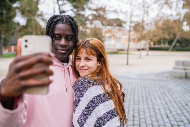 Parkta güneşli bir günün tadını çıkaran genç ve çok ırklı bir çift kucaklaşırken ve gülümserken neşeli bir selfie çekiyorlar.