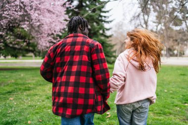 İlkbaharda el ele tutuşup parkta yürüyen ve birlikte romantik bir anın tadını çıkaran çok kültürlü genç bir çift.