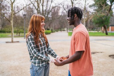 Genç, çok ırklı bir çift parkta el ele tutuşuyor ve gülümsüyor. Birlikte romantik bir anın tadını çıkarıyorlar.