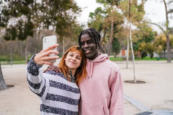 Canlı bir parkta akıllı bir telefonla selfie çekerken gülümseyen mutlu çok ırklı çift, doğada birlikte vakit geçirmenin keyfini çıkarıyorlar.