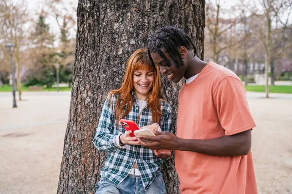 Parkta ağaç gövdesine yaslanırken akıllı telefonlar kullanan ve gülümseyen çok ırklı mutlu çift.