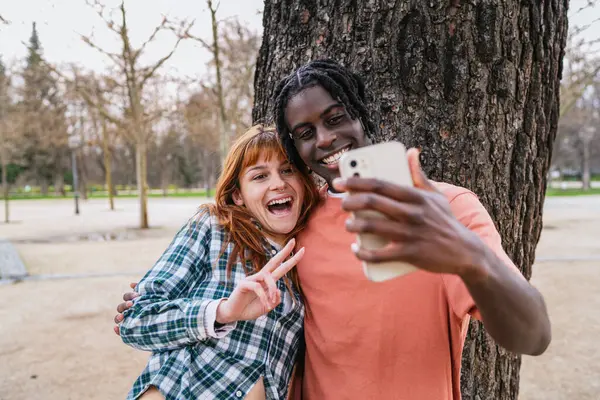 Genç, çok ırklı bir çift gülümseyip selfie çekerken barış işareti yapıyor, canlı bir parkta neşeli bir anın tadını çıkarıyorlar.