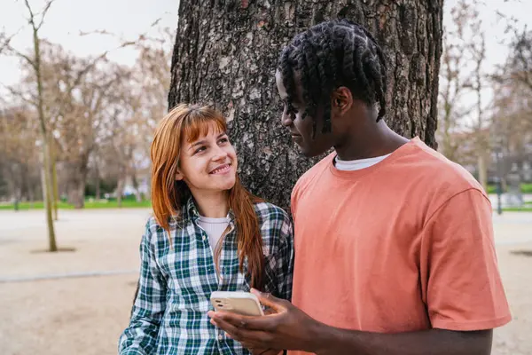 Genç, çok ırklı bir çift gülümsüyor ve akıllı bir telefon kullanıyor. Bu arada yeşil alanlarla dolu canlı bir parkta birlikteliklerinin tadını çıkarıyorlar.