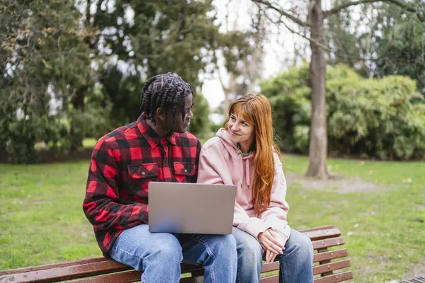 Parktaki bankta dizüstü bilgisayar kullanan ve birbirlerine bakıp gülümseyen çok kültürlü genç çift.