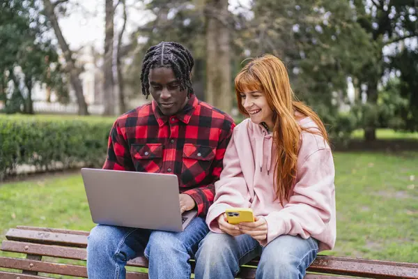 Genç, çok ırklı bir çift, parktaki bankta oturuyor. Çevrelerindeki doğanın güzelliğinin tadını çıkarırken dizüstü bilgisayar ve akıllı telefon kullanıyorlar.