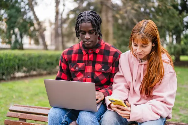 Genç çok ırklı öğrenciler bir üniversite projesinde dizüstü bilgisayar ve akıllı telefon kullanarak bir parkta bankta oturuyorlar.