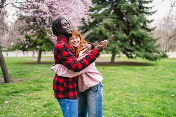 Genç, çok ırklı bir çift bahar aylarında parkta sarılırken akıllı telefonlarıyla selfie çekiyor.