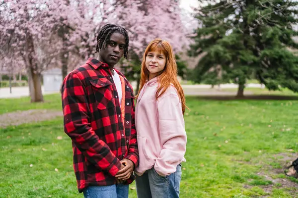 Parkta güneşli bir günün tadını çıkaran çok kültürlü genç bir çift. İlkbaharda açan kiraz ağaçları ve canlı çiçeklerle çevrili.