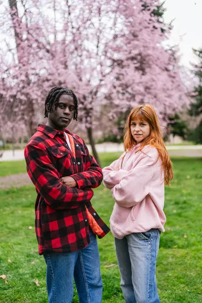 Genç, çok ırklı bir çift parkta çimlerin üzerinde dikilip arka planda çiçek açan ağaçlarla kollarını kavuşturuyor.