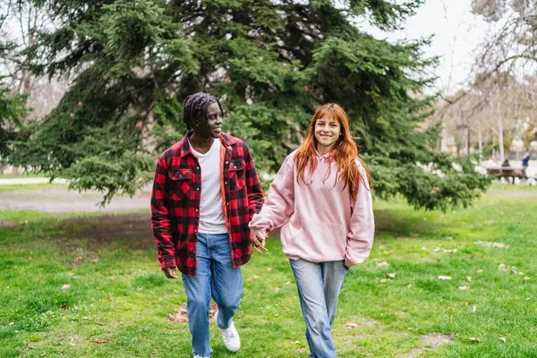 Genç, çok ırklı bir çift, canlı yeşil bir parkta yürürken el ele tutuşuyor, birlikte vakit geçiriyorlar ve neşeli anlarını paylaşıyorlar.