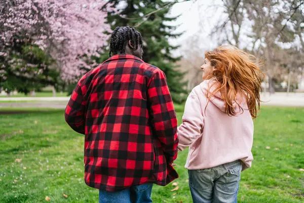 İlkbaharda el ele tutuşup parkta yürüyen ve birlikte romantik bir anın tadını çıkaran çok kültürlü genç bir çift.