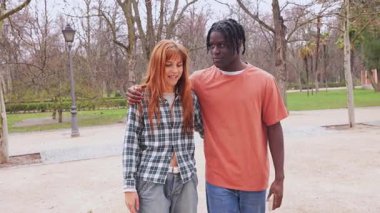 Irklar arası çift birlikte yürüyor, güneşli park manzarasının tadını çıkarırken samimi bir sohbet ediyorlar, gerçek bir bağ ve sevgi gösteriyorlar.