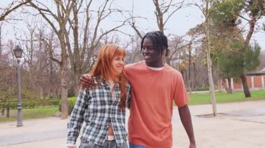 Irklar arası genç çift kol kola yürüyorlar, neşeli sohbetler ediyorlar ve güneşli park yolunda yürürken sevgi dolu ilişkiler kuruyorlar.