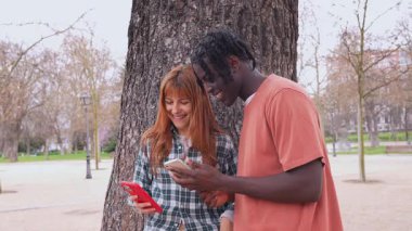 Irklar arası genç çift akıllı telefon anlarını paylaşıyor, park yeşillikleri arasında dijital bağlantının keyfini çıkarıyorlar.