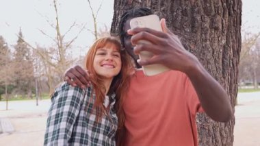 Genç, çok ırklı bir çift, birlikte güneşli bir parkın keyfini çıkarırken, eğlenceli bir şekilde selfie çekiyor romantik anları yakalıyor.