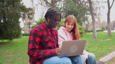 Uzaktan bilgisayarla çalışan genç bir çift, yeşil alanlarla çevrili park bankında birlikte oturuyorlar, dijital çalışma alanını sevgi ve bağlantıyla paylaşıyorlar.